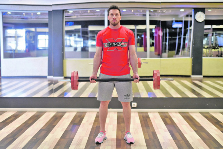 VEŽBA 4 (STOJEĆI BICEPS PREGIB) - korak 1 - Dok smo u startnom položaju, noge su u širini ramena. Opterećenje je u rukama, a šipku hvatamo takozvanim pothvatom, odnosno odozdo. Šake su u pothvatu.